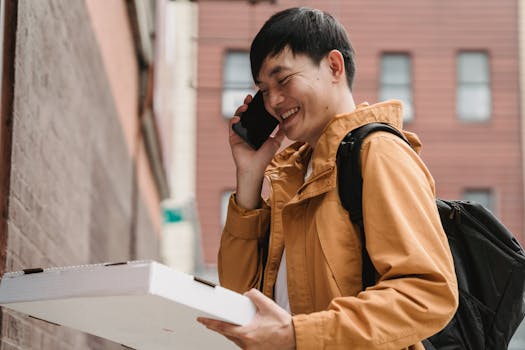 Asian male delivery person with pizza box, smiling while on a call in a city street.