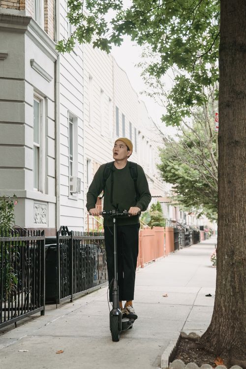 Free A fashionable man in a beanie rides an electric scooter on a tree-lined city sidewalk. Stock Photo