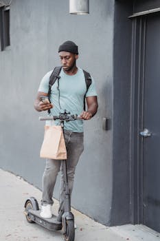 A delivery man with a backpack checks his phone while riding an electric scooter outdoors.