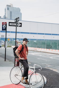 A delivery cyclist in urban attire pauses on a city street corner, with tall buildings in the background.
