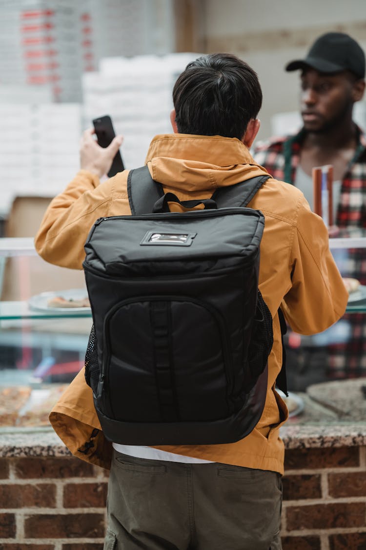 Backpack On The Back Of A Man Showing His Phone To An Employee
