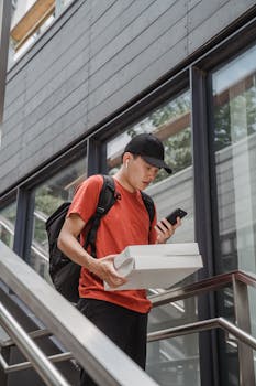 A courier navigating city stairs while checking a smartphone, holding delivery boxes.