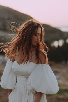 A woman in a white off-shoulder dress at sunset with flowing hair outdoors.