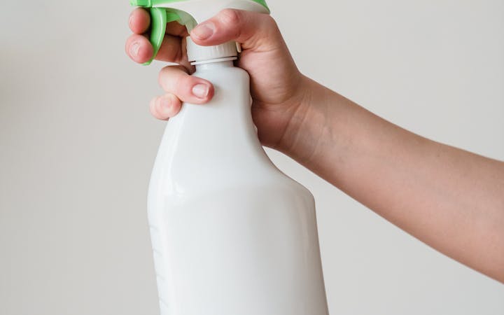 Close-up of a hand holding a white spray bottle with a green nozzle on a neutral background.