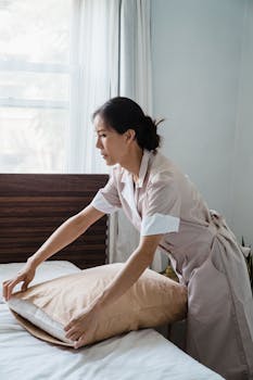 Asian woman in a tan uniform arranging a pillow in a bright, sunlit bedroom.