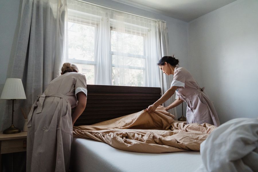 Two cleaners making a bed with fresh linens in a bright, sunlit bedroom