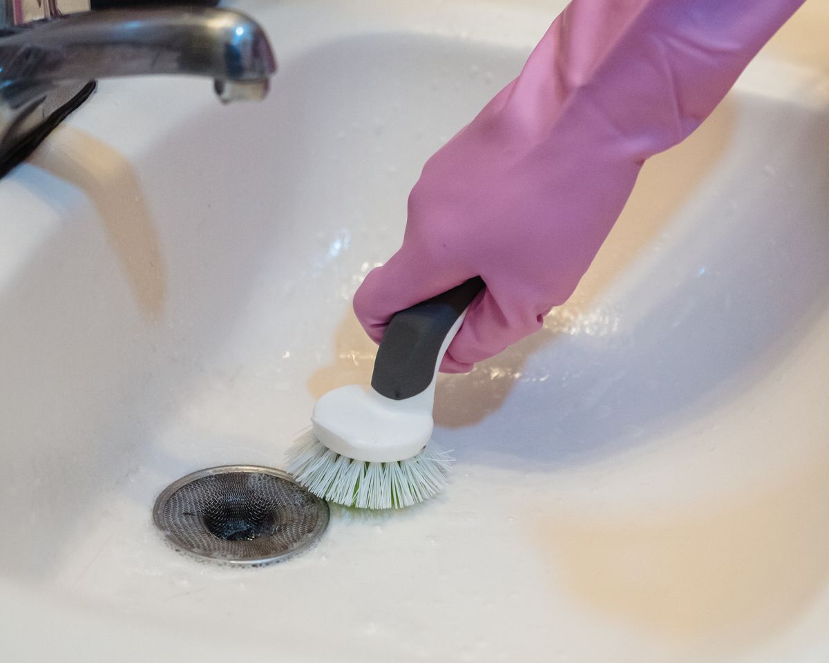 Close-up of a hand in pink gloves cleaning a sink with a brush and faucet visible