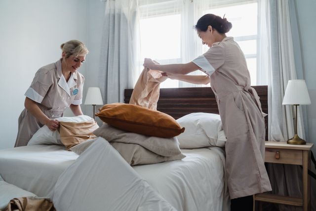 Two housekeepers in uniform fixing a hotel room bed with fresh linen and pillows. Related image for Hotel Operations: Trends Shaping Hotels in 2026.