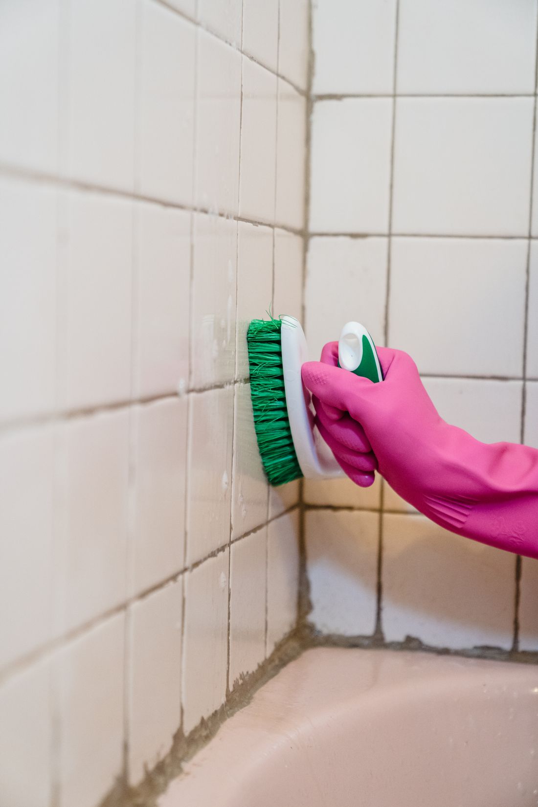 Gloved hand scrubbing grout lines between white wall tiles
