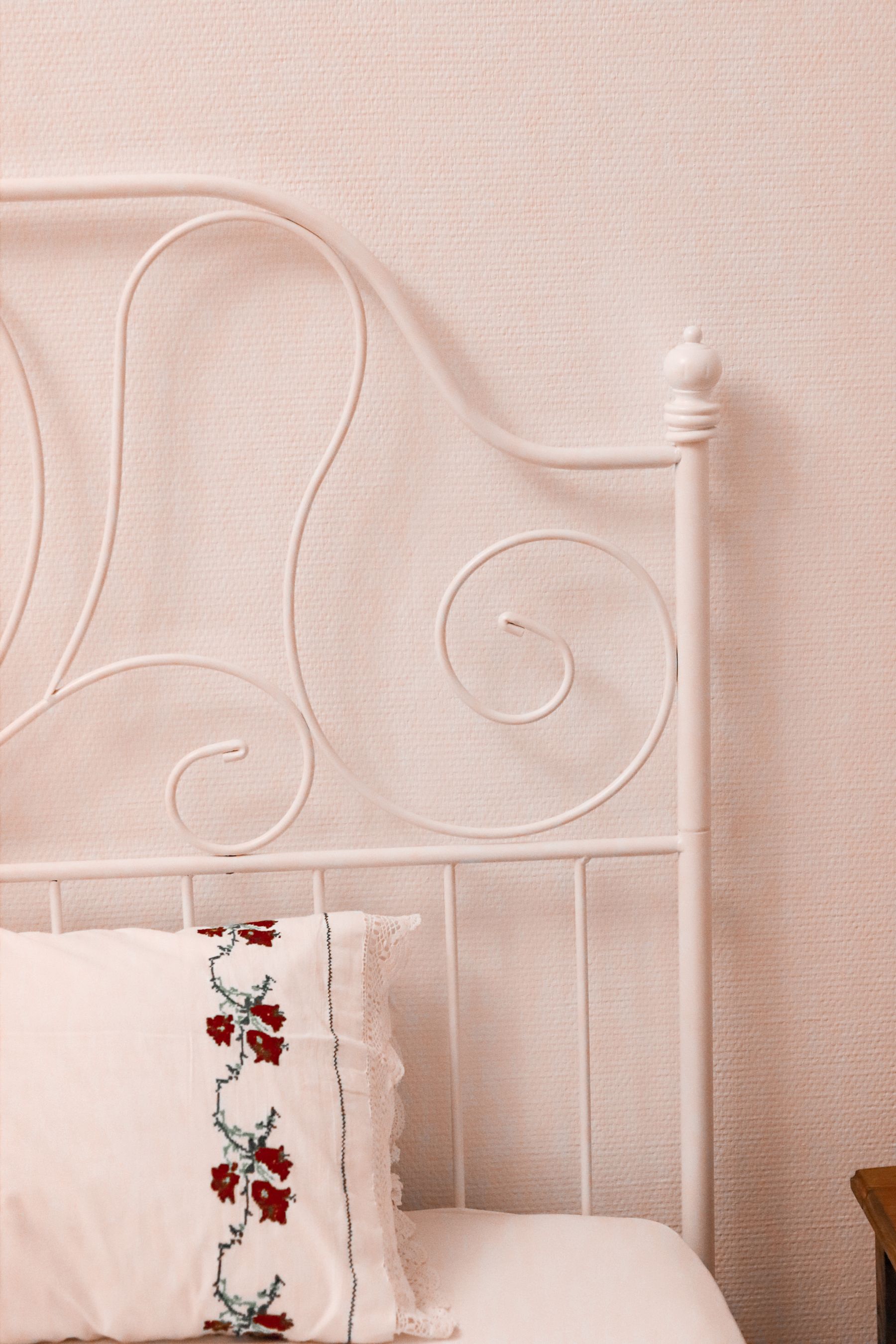 White iron headboard with a pastel floral embroidered pillow against a pale pink wall