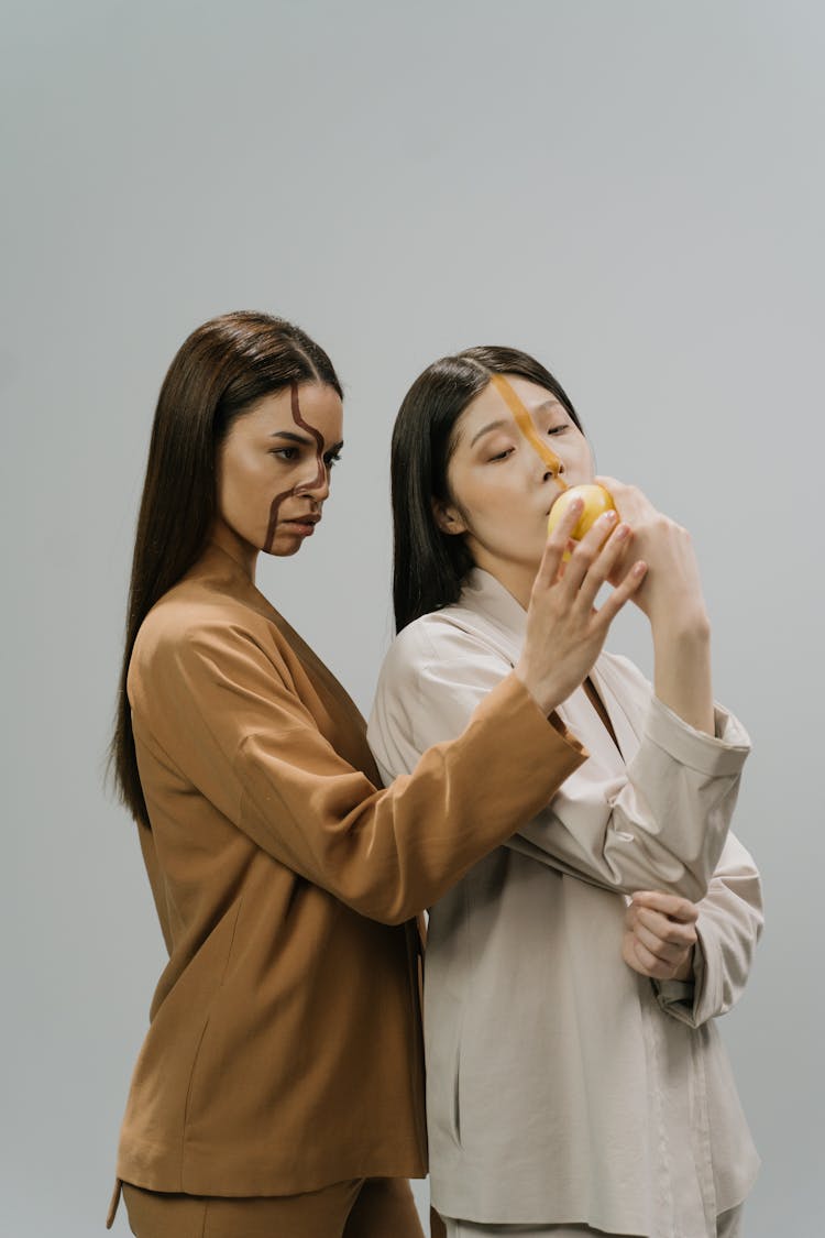 A Woman Standing Behind Another Woman Biting A Fruit