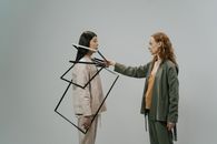 Young female model in green costume and beige top putting empty photo frames on shoulders of other female model wearing light beige jacket and trousers