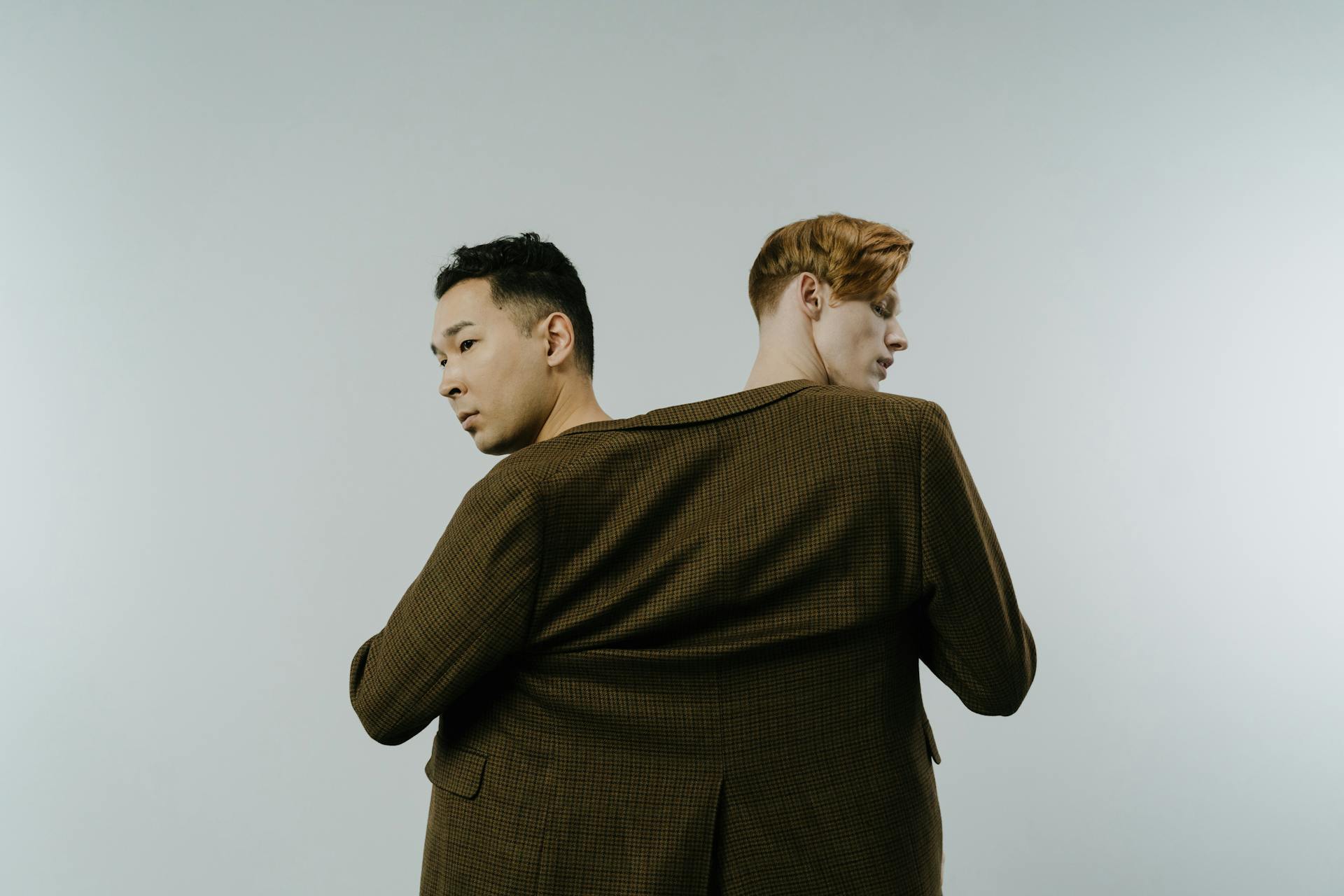 Two men creatively sharing a single large brown blazer, facing opposite directions against a minimalist backdrop.