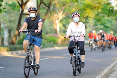 Free Asian couple cycling with masks and helmets on a sunny day in Jakarta. Stock Photo