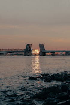 Peaceful sunrise with drawbridge raised over calm river water reflecting warm colors.