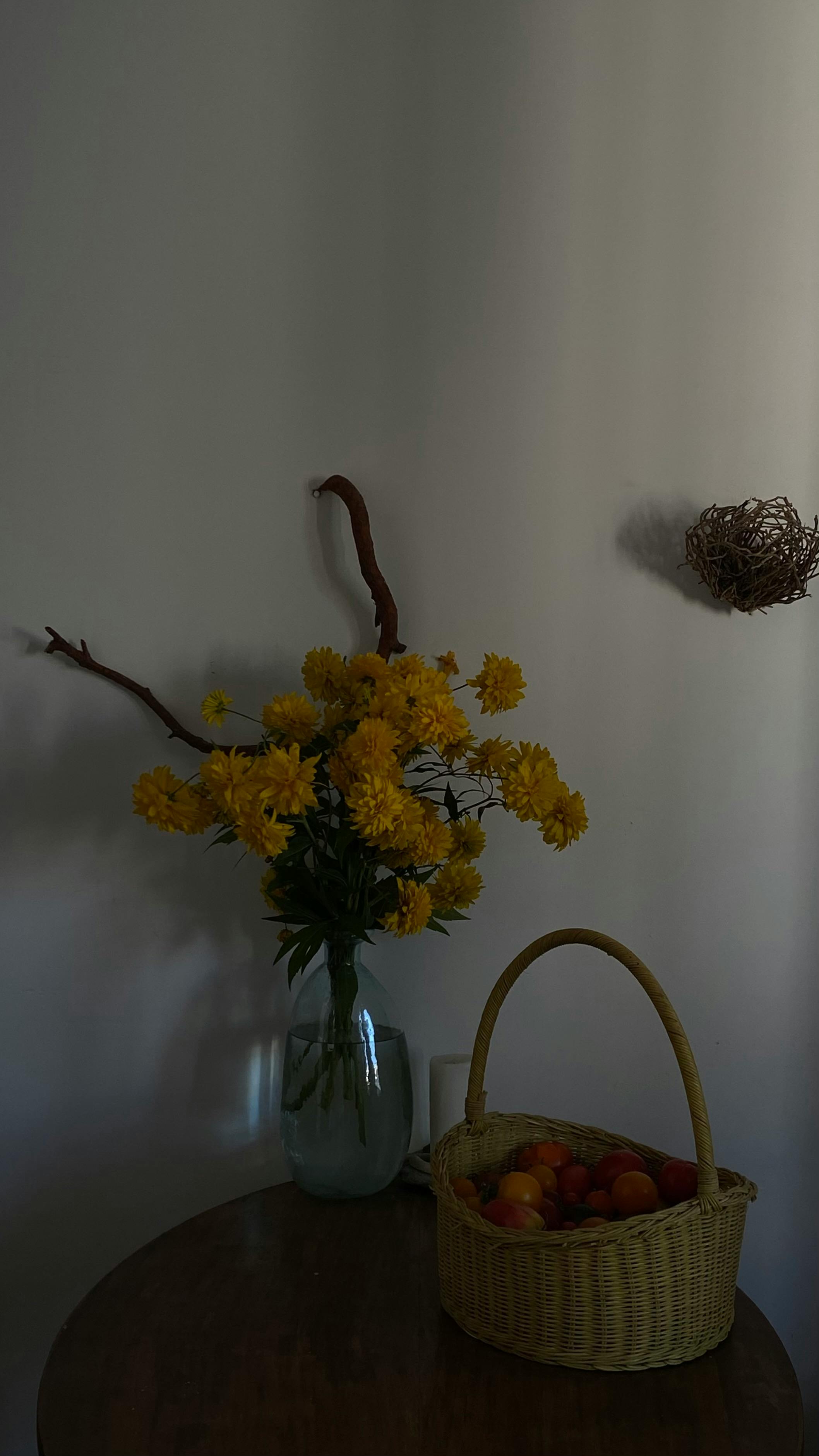 A serene still life featuring a vase with yellow flowers and a fruit-filled basket on a table.
