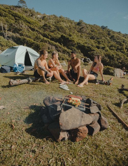 Fotomural Amigos se reúnen en un camping escénico asando comida cerca de una tienda, abrazando la aventura al aire libre.