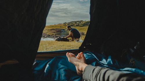 Free Relaxing inside a tent with a view of a camper preparing outdoors by a serene coastal landscape. Stock Photo