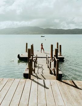 A woman in swimwear strolls along a picturesque wooden dock on a sunlit lake surrounded by mountains.