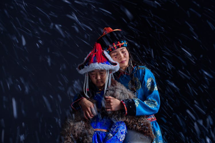 Woman And Child In Traditional Clothing Sitting Outside During A Snowfall 