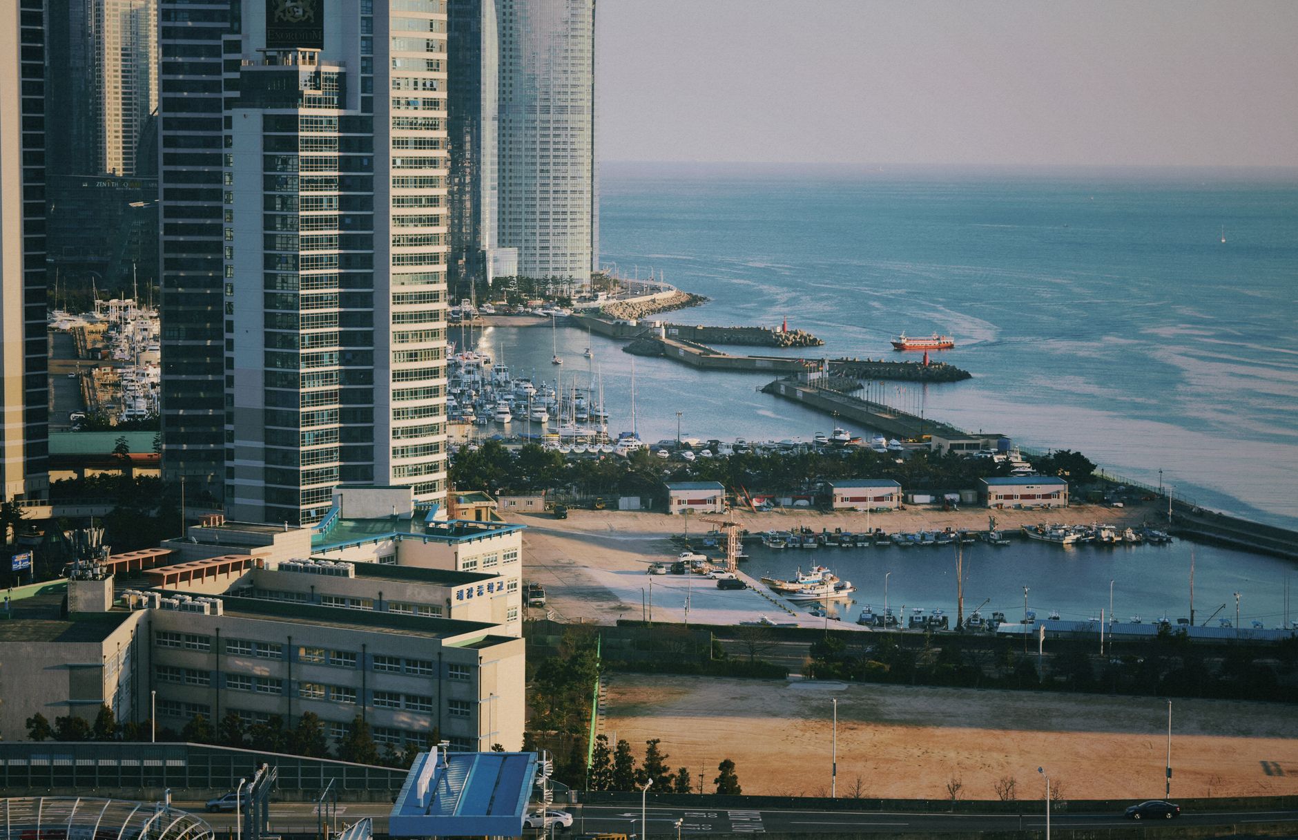잔잔한 해안선 근처의 마리나에서 촬영한 고층 빌딩의 공중 사진으로, 요트와 도시 풍경이 보입니다.