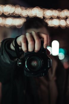 Close-up of a person holding a DSLR camera with blurred bokeh lights in background.