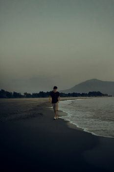 A lone individual strolls along a quiet beach with a mountainous backdrop, waves gently lapping at the shore.