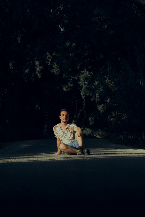 Free A man in casual attire sitting on a shadowy road surrounded by greenery, creating a peaceful scene. Stock Photo