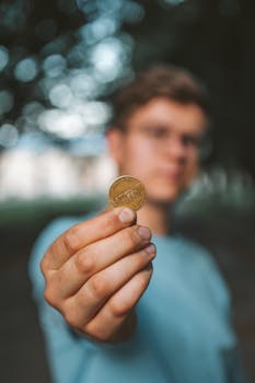 A young adult holds a coin up close with a blurred natural background, focusing on finance concept.
