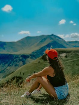 A woman sits on a grassy hillside wearing a red cap, enjoying a view of mountains under a clear blue sky.