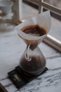 A Chemex brewing coffee on a digital scale by a window, evoking warmth and relaxation.