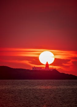 Silhouetted lighthouse against a vibrant orange sunset over the sea, creating a dramatic and serene scene.