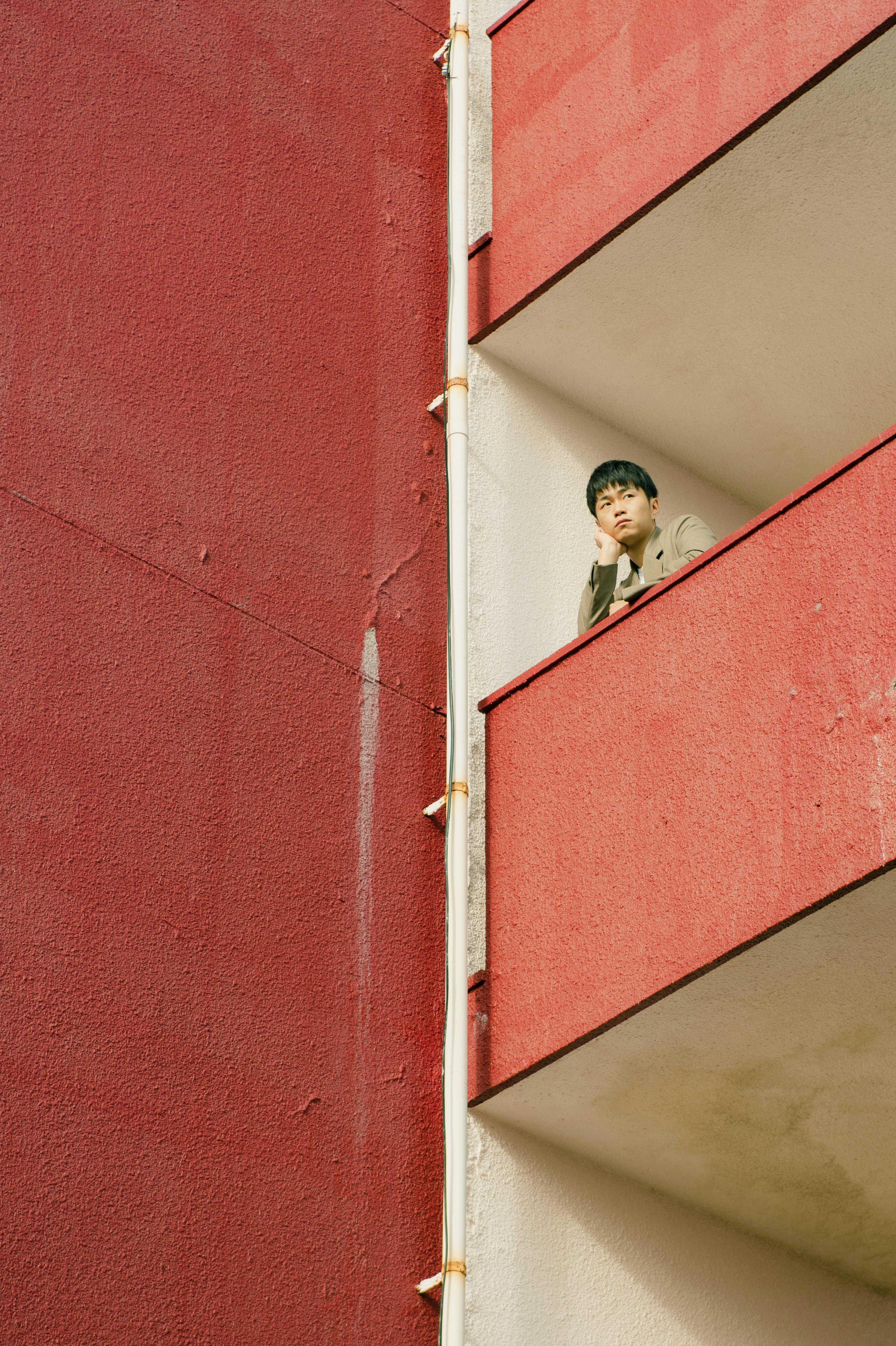 An individual stands thoughtfully on a red building's balcony, symbolizing city isolation.