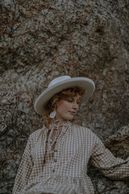 Caucasian woman in checkered dress and hat, thoughtful outdoors against rocky background.