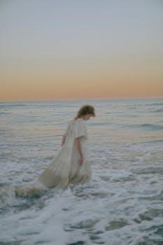Woman in a white dress walking through ocean waves at sunset, creating a serene atmosphere.