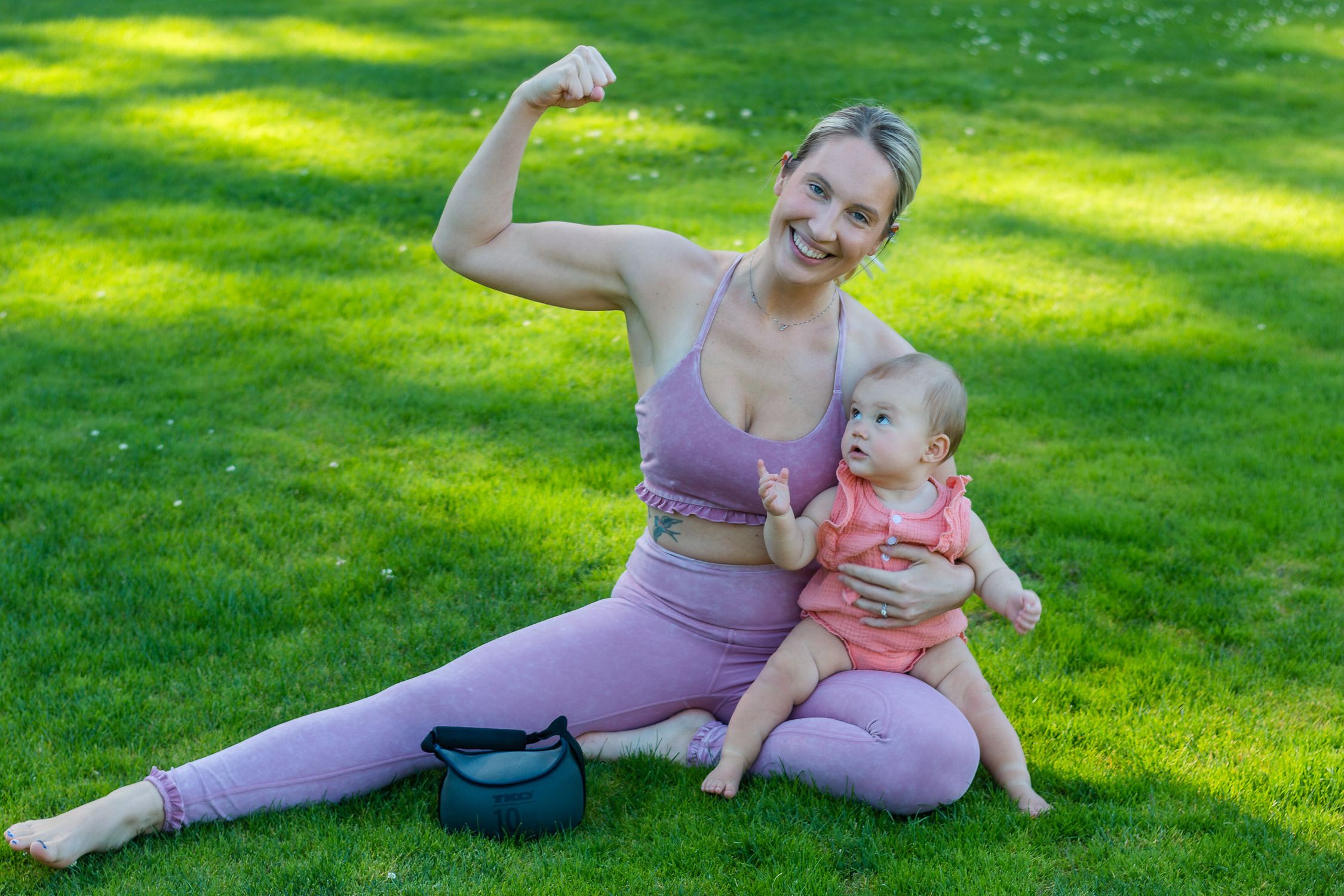 Uma mãe sorridente flexiona os músculos brincando enquanto sentada com seu bebê em um gramado ensolarado.