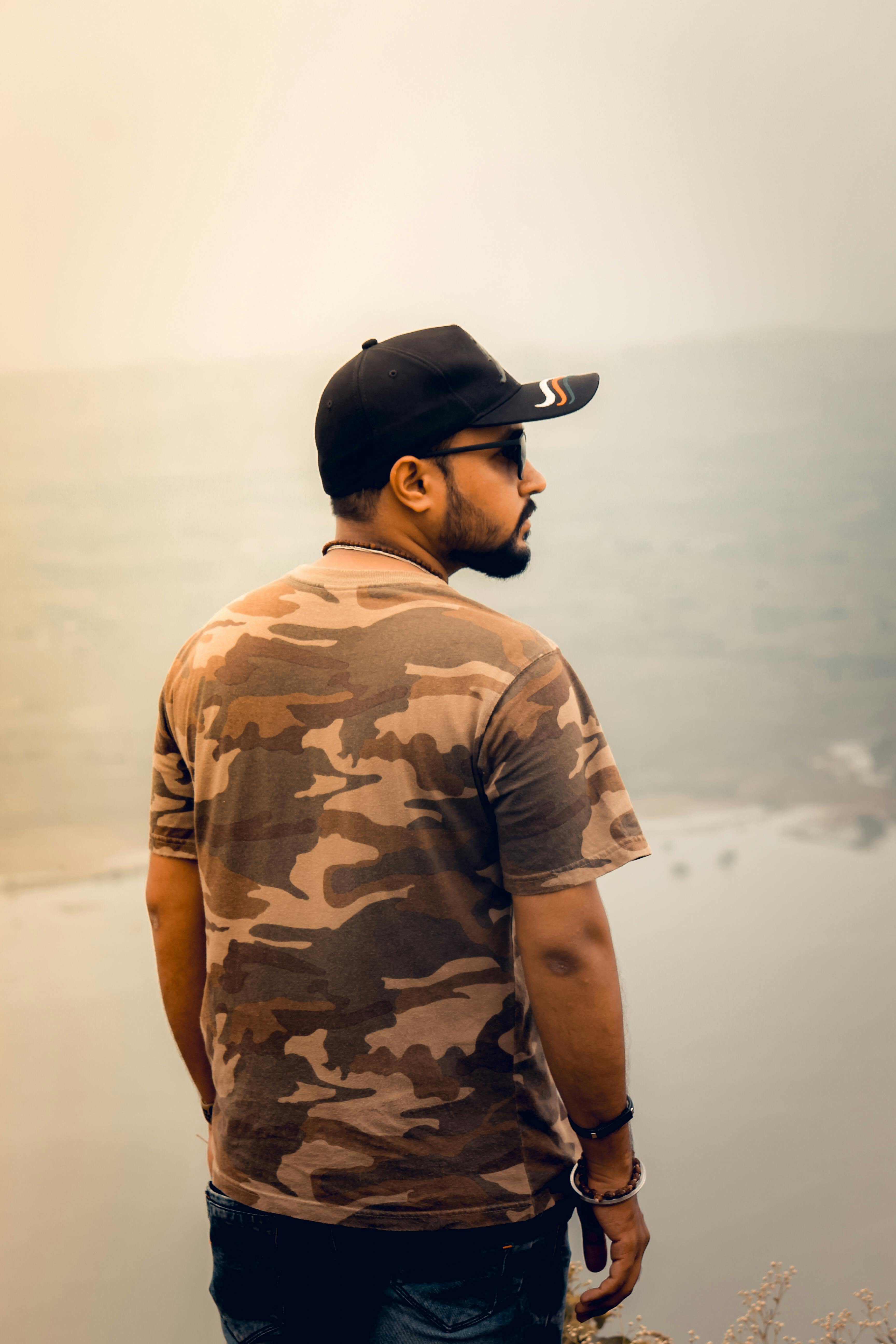 A man wearing a cap and camouflage shirt stands by a serene lake with soft lighting.