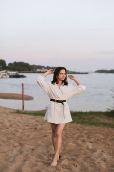 A woman in a white dress walks barefoot on a sandy riverbank, enjoying a serene summer evening.