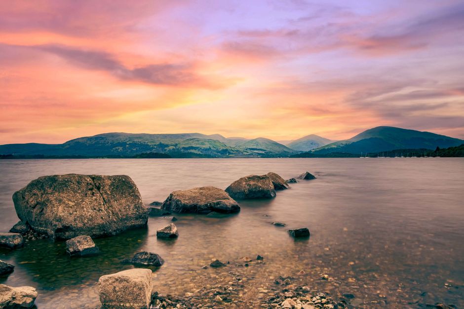 Peaceful sunset view over Loch Lomond with rocky shoreline and distant hills in Scotland.