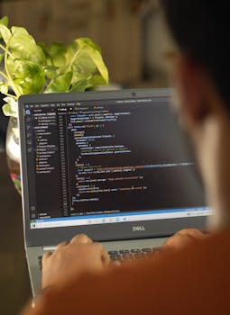 Person focused on coding with laptop and plant in view, highlighting a modern work environment.