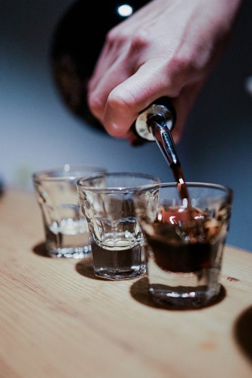 Person Pouring Brown Drink in Clear Shot Glasses