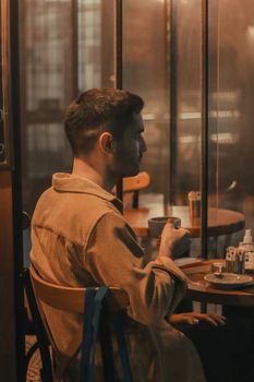 Man seated in a cozy café, wearing velvet shirt, enjoying a warm drink at night.