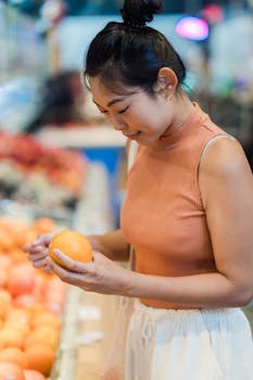 Asian woman selecting fresh oranges in a grocery store aisle, shopping for fruits.