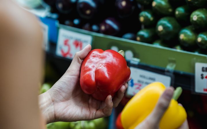 A woman chooses between red and yellow bell peppers at a supermarket shelf.
