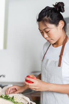 Asian woman in kitchen holding a fresh red apple, wearing an apron.