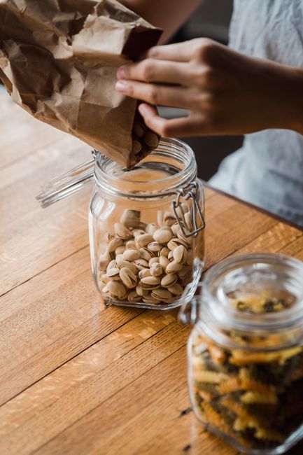 Hands pouring pistachios from brown paper bag into a glass jar on a wooden table. - Effective Storage Methods for Dry Fruits