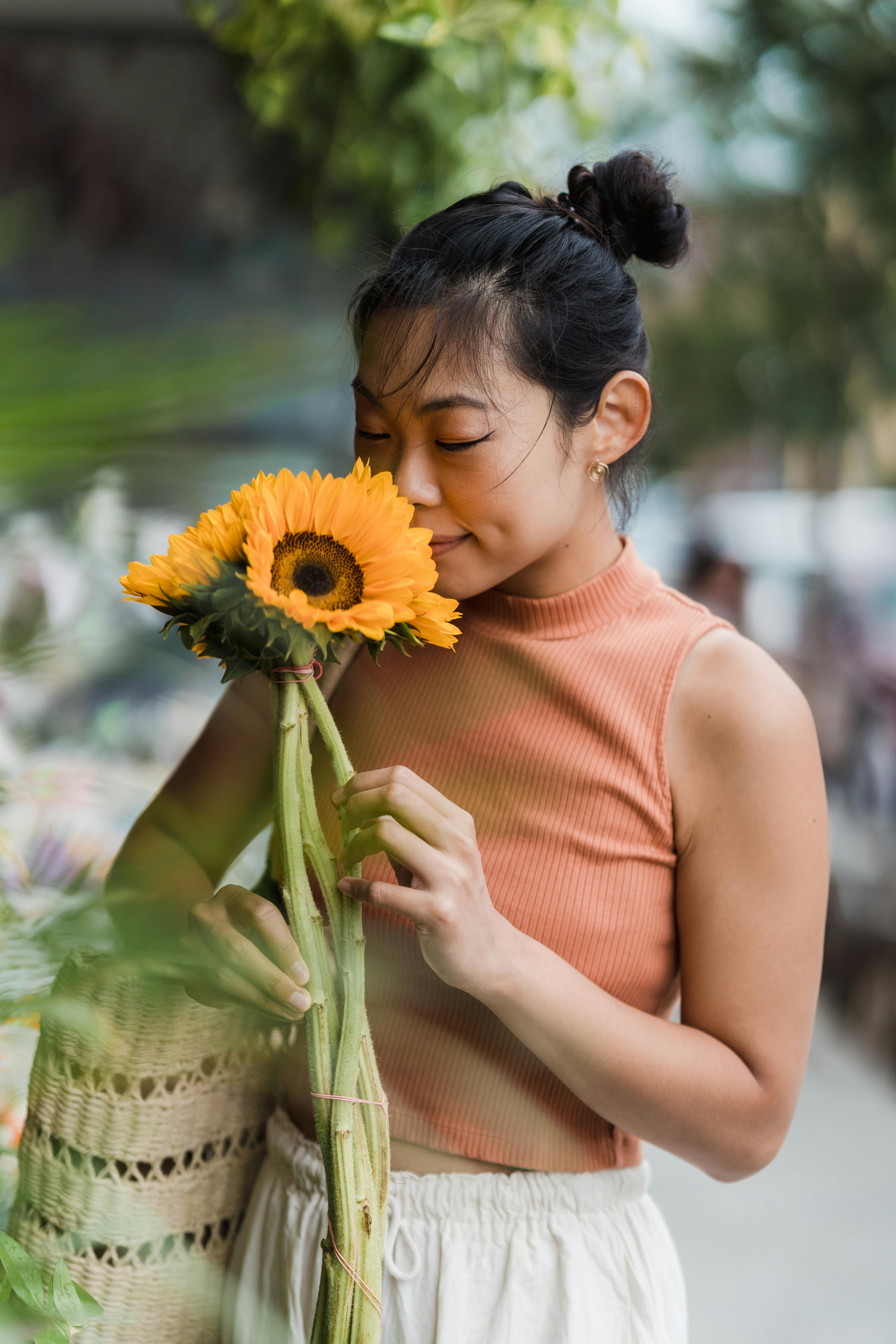 Asian woman holding and smelling a sunflower, enjoying its fragrance outdoors.
