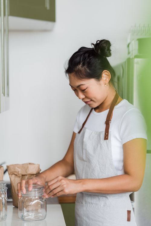 Free Asian woman wearing apron holding a glass jar in a kitchen setting with a focus on food preparation. Stock Photo