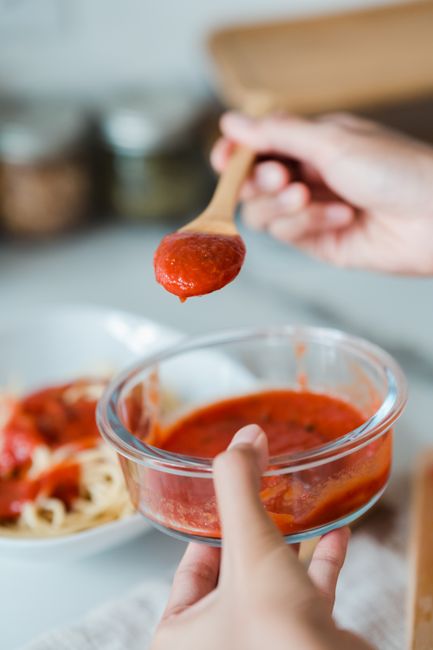 Hand holding a small glass jar of red pasta sauce over a bowl