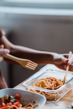 Close-up of hands using wooden forks to eat spaghetti from a glass container indoors.